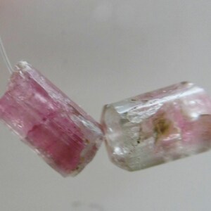 May include: Two pink and clear tourmaline gemstone beads. The cylindrical beads are strung on a thin, clear string. The gemstones have a translucent appearance with pink and clear color variations.