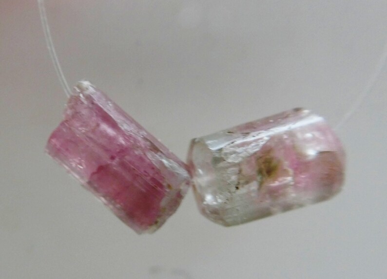May include: Two pink and clear tourmaline gemstone beads. The cylindrical beads are strung on a thin, clear string. The gemstones have a translucent appearance with pink and clear color variations.