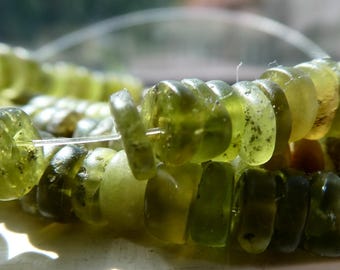 Peut inclure: Gros plan sur un fil de perles de pierres précieuses vert olive. Les perles sont de forme irrégulière et translucides, avec de petites inclusions sombres. Les perles sont enfilées sur un cordon fin et transparent.