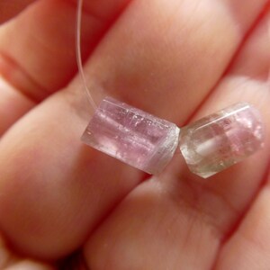 May include: Two translucent, pink and clear tourmaline gemstone beads on a white string. The beads are rectangular with rounded edges. The gemstones are held in a hand.