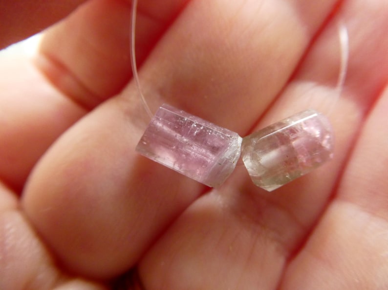 May include: Two translucent, pink and clear tourmaline gemstone beads on a white string. The beads are rectangular with rounded edges. The gemstones are held in a hand.