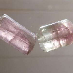May include: Two translucent, rectangular tourmaline beads, one pink and the other with pink and green hues, are strung on a thin, clear cord. The gemstones are polished and have visible internal structures.