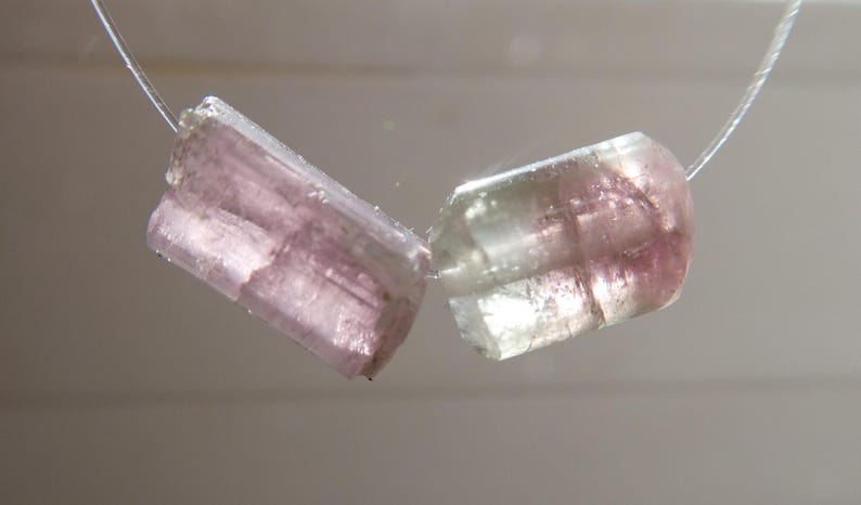 May include: Two translucent, rectangular tourmaline beads, one pink and the other with pink and green hues, are strung on a thin, clear cord. The gemstones are polished and have visible internal structures.