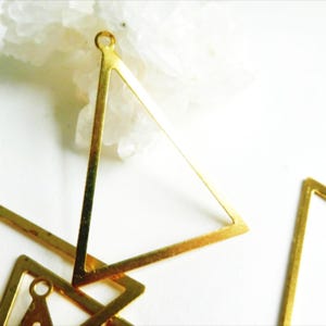 May include: Several gold-colored, geometric triangle charms with small loops for jewelry making. The charms are arranged on a white surface with a white, crystalline object in the background.