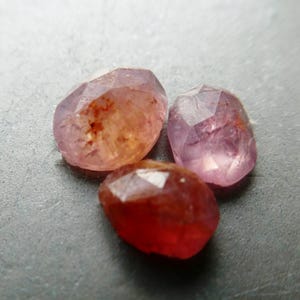May include: Three faceted gemstones in shades of pink and red. The gems have irregular shapes and a polished, reflective surface. The stones are arranged on a gray surface, with varying tones of pink and red.
