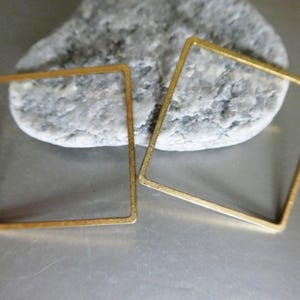 May include: Two gold-colored square earring components. The open-frame squares are displayed on a gray stone surface. The earrings are made of a thin metal and have a simple, geometric design.