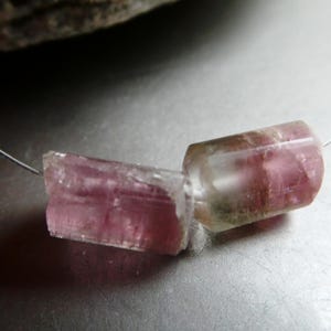 May include: A delicate necklace featuring two raw, pink tourmaline gemstone beads. The beads are cylindrical and translucent, with visible internal textures and color variations. The necklace is strung on a thin, silver-colored wire.