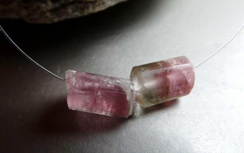 May include: A delicate necklace featuring two raw, pink tourmaline gemstone beads. The beads are cylindrical and translucent, with visible internal textures and color variations. The necklace is strung on a thin, silver-colored wire.