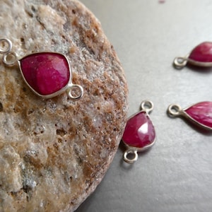 May include: Several ruby gemstone pendants with silver settings are displayed. The pendants have a vibrant red hue and various shapes, including teardrop and square-cut designs. Each pendant has a small loop for attaching to a chain or other jewelry component.