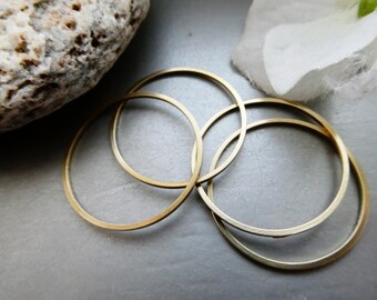 May include: Four gold-colored hoop earrings, varying in size, are arranged on a gray surface. The hoops are thin and circular, with a polished finish. A light-colored rock and a white fabric are in the background.
