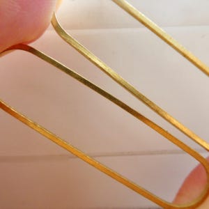 May include: Close-up of two gold-colored rectangular earrings. The earrings are held between fingers, showcasing their simple, elegant design. The earrings have a smooth, polished finish, reflecting light. The overall aesthetic is minimalist and modern, suitable for everyday wear.