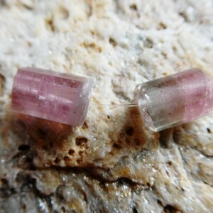 May include: Two translucent, pink tourmaline beads are strung on a thin, clear cord. The cylindrical beads have a slightly rectangular shape and are set against a textured, beige background. These beads are ideal for jewelry making.