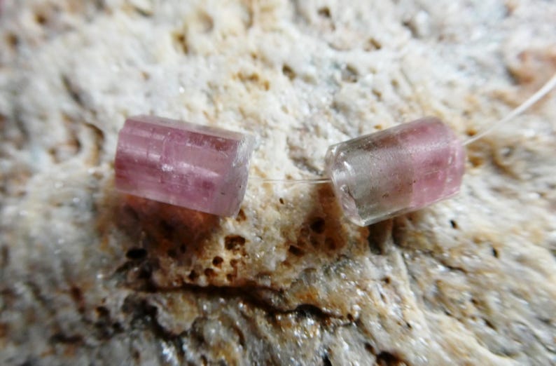 May include: Two translucent, pink tourmaline beads are strung on a thin, clear cord. The cylindrical beads have a slightly rectangular shape and are set against a textured, beige background. These beads are ideal for jewelry making.