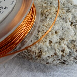 May include: A clear plastic spool containing bright orange copper wire, approximately 13.72 metres, is shown. The wire is coiled and sits on a textured, light grey stone. The spool has text on it.