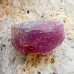 May include: A faceted, raw ruby gemstone, displaying a deep pink hue. The gem is set against a textured, light-colored stone surface. The ruby's crystalline structure is visible, showcasing its natural form.