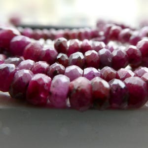 May include: Close-up of a strand of faceted, deep red ruby beads. The beads are irregularly shaped and have a polished, reflective surface. The beads are strung together, forming a necklace or bracelet.
