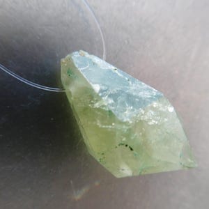 May include: A light green crystal pendant with a clear string. The crystal has a rough, faceted surface with some darker green inclusions. The pendant is a pale green color.
