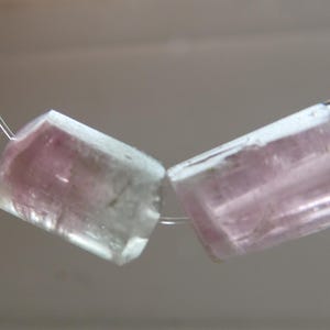 May include: Two rectangular, translucent gemstones with pink and clear hues are strung on a thin, silver-colored wire. The gemstones are cut with angled edges, and the pink color is concentrated in the center of each stone.