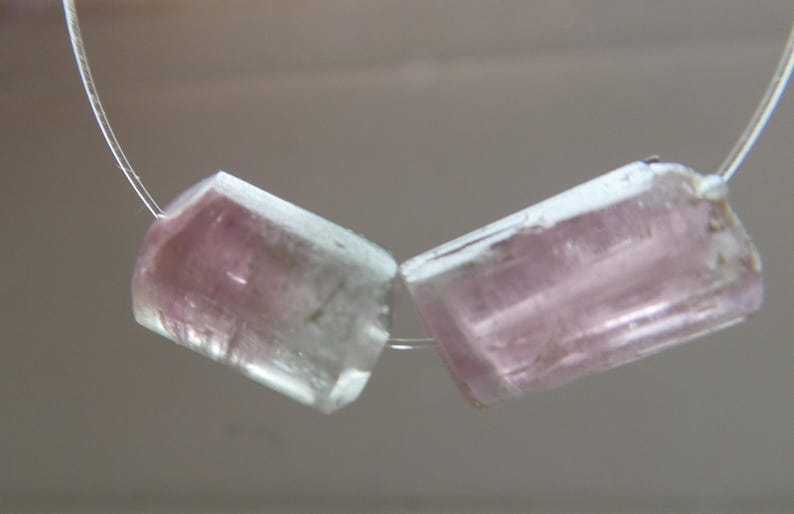 May include: Two rectangular, translucent gemstones with pink and clear hues are strung on a thin, silver-colored wire. The gemstones are cut with angled edges, and the pink color is concentrated in the center of each stone.