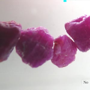 May include: Four raw, unpolished ruby gemstones with a deep magenta-pink hue, strung together on a thin, clear filament. The stones have irregular, jagged shapes and textured surfaces. The text "No 2" is printed in the bottom right corner.