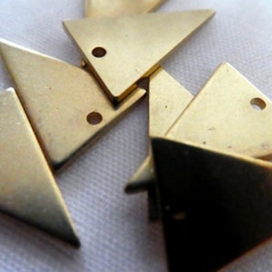 Op de afbeelding: Een groep van acht gouden driehoekige metalen bedels met een gat in elke hoek. De bedels liggen verspreid op een wit oppervlak.