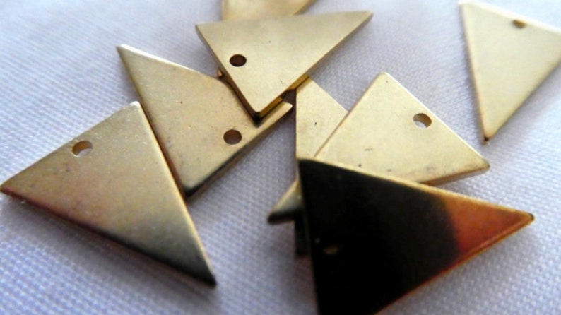 Op de afbeelding: Een groep van acht gouden driehoekige metalen bedels met een gat in elke hoek. De bedels liggen verspreid op een wit oppervlak.