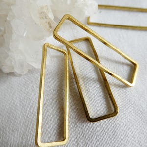 May include: Close-up of several gold-colored rectangular metal earring components. The open rectangles are arranged on a white surface, with a piece of white crystal in the background. The earrings have a simple, geometric design.