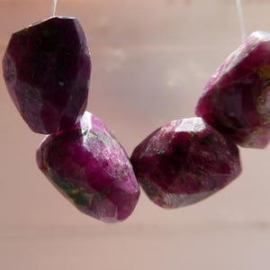 May include: Four faceted, deep red-purple ruby gemstones strung on a white thread. The gemstones have a polished, irregular shape and are suspended against a soft, neutral background. The gems are a rich, saturated color.