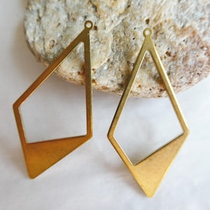 May include: Two brass-colored geometric earring pendants. One is a diamond shape, and the other is a modified diamond shape. Both have a small loop at the top for attachment. The pendants are displayed on a light-colored, textured surface.