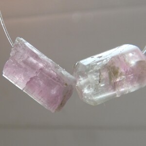 May include: Two raw, translucent tourmaline crystals with pink and clear hues are strung on a thin, clear cord. The crystals are rectangular with rough, natural edges, showcasing their unique, organic form.