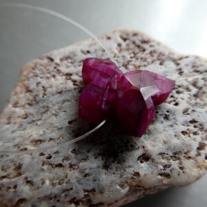 May include: A deep magenta, faceted gemstone carved into the shape of a butterfly. The stone features a clear, thin filament thread passing through its center, allowing it to be suspended or strung as a decorative bead or jewelry component.