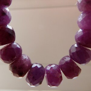 May include: A strand of faceted, deep magenta-colored gemstone beads. The beads are irregularly shaped and strung together, creating a curved line. The gemstones have a polished, reflective surface.