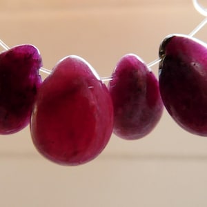 May include: Four polished, teardrop-shaped, deep magenta gemstones strung on a thin, clear cord. The gems have a glossy finish and vary slightly in size. The image is set against a soft, neutral background.