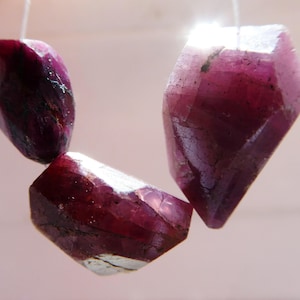 May include: Three faceted ruby gemstones suspended by thin white threads. The gemstones are a deep, rich red with variations in colour and texture. The light catches the facets, creating highlights and shadows.