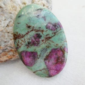 Peut inclure: Une pierre précieuse polie de forme ovale présentant une surface lisse avec un motif marbré composé d'inclusions vert menthe et magenta vif, accentué par de fines veines sombres en forme de toile sur toute la face arrondie et polie.