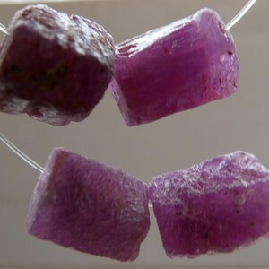 May include: Four rough-cut, deep pink gemstones strung on a thin, clear wire. The stones are irregularly shaped with a textured surface. The text "No 1" and "No 2" are visible. These appear to be raw ruby gemstones.