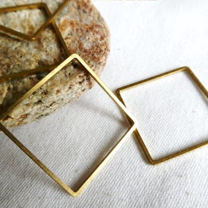 May include: Several open square-shaped brass metal frames. One frame is on a textured rock, and two are on a white surface. The frames are a warm gold color and have a simple, geometric design. The frames are likely for jewelry making.