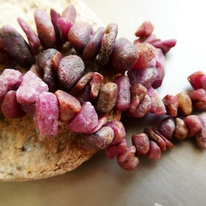 May include: A strand of raw ruby gemstone beads in varying shades of pink and brown. The irregular, tumbled stones are strung together, creating a natural and earthy aesthetic. The beads are displayed on a light-colored, textured surface.