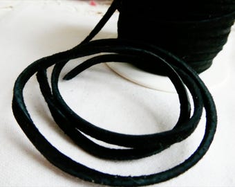 May include: A spool of black suede cord with a length of the cord unwound in the foreground. The cord is a deep black color and appears soft to the touch. The cord is likely used for jewelry making or crafting.