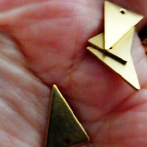 Op de afbeelding: Drie gouden driehoekige metalen bedels met gaten om te rijgen. De bedels zijn op elkaar gestapeld.