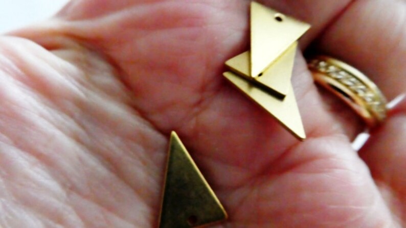 Op de afbeelding: Drie gouden driehoekige metalen bedels met gaten om te rijgen. De bedels zijn op elkaar gestapeld.