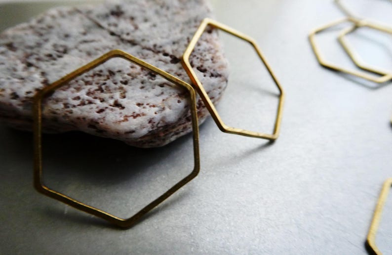 Peut inclure: Plusieurs cadres hexagonaux en m&eacute;tal dor&eacute; fin avec une finition lisse. Les formes g&eacute;om&eacute;triques sont dispos&eacute;es avec un cadre reposant partiellement sur une pierre textur&eacute;e de couleur claire, tandis que d'autres cadres identiques sont dispers&eacute;s.