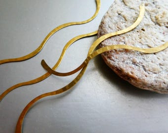 Könnte beinhalten: Nahaufnahme von goldfarbenen, wellenförmigen Metallschmuckstücken. Der Schmuck ist auf einem hellen Stein mit grauem Hintergrund angeordnet. Das Metall hat eine gehämmerten Textur, und das Design ist abstrakt und fließend.