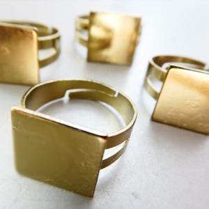 May include: Four gold-toned metal rings featuring a flat, square-shaped top surface. Each ring has an adjustable band design with a split shank, providing a metallic finish that reflects light across the smooth, square face of the jewelry piece.