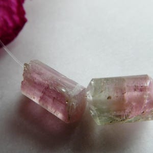 May include: Two raw, pink and green tourmaline gemstone beads on a thin, clear string. The gemstones have a translucent appearance with visible internal structures. A blurred pink petal is in the background.