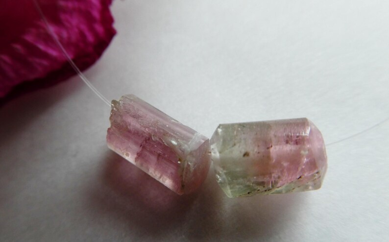 May include: Two raw, pink and green tourmaline gemstone beads on a thin, clear string. The gemstones have a translucent appearance with visible internal structures. A blurred pink petal is in the background.