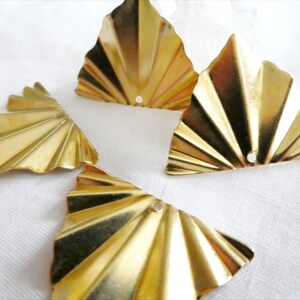 May include: Four gold-colored, fan-shaped metal components with a ridged design. Each piece has a small hole near the center. The objects are arranged on a white, textured surface, creating a visually appealing display.
