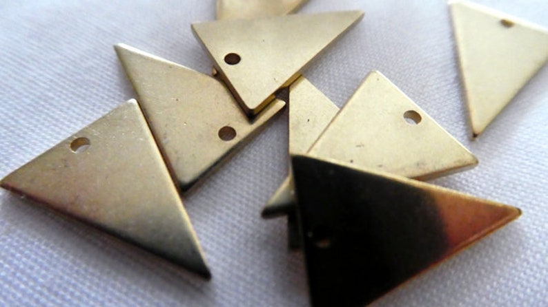 Op de afbeelding: Een groep van zes gouden driehoekige metalen bedels met een gat in elke hoek. De bedels zijn willekeurig gerangschikt op een wit oppervlak.