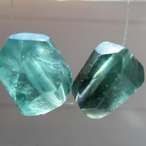 May include: Two faceted, translucent teal-colored beads with irregular, geometric shapes. Each bead features multiple flat surfaces that catch the light, creating internal reflections and depth. The beads are strung on a thin, clear filament.