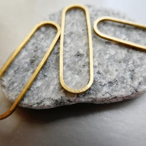 May include: Close-up of three gold-colored, elongated oval earrings. The earrings are placed on a gray and white stone surface. The earrings have a simple, minimalist design, with a smooth, polished finish. The overall aesthetic is modern and elegant.